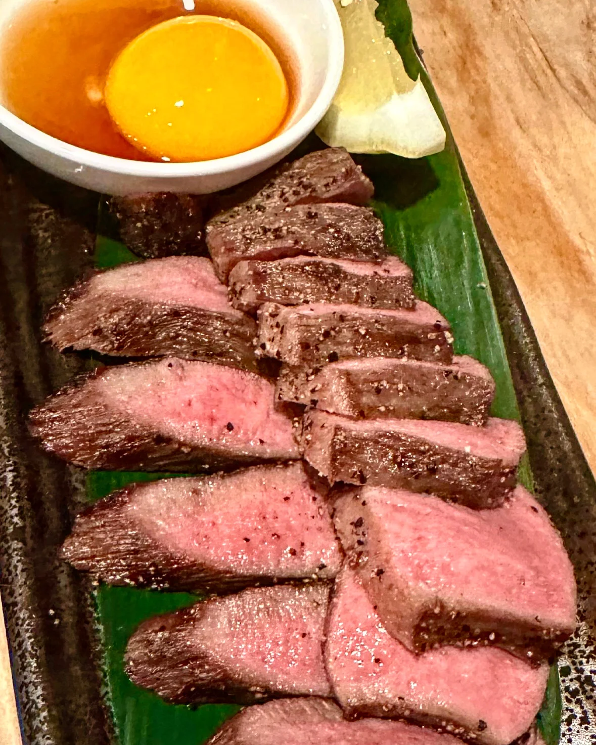 Slices of seared beef on a dark plate beside a raw egg yolk and dipping sauce.