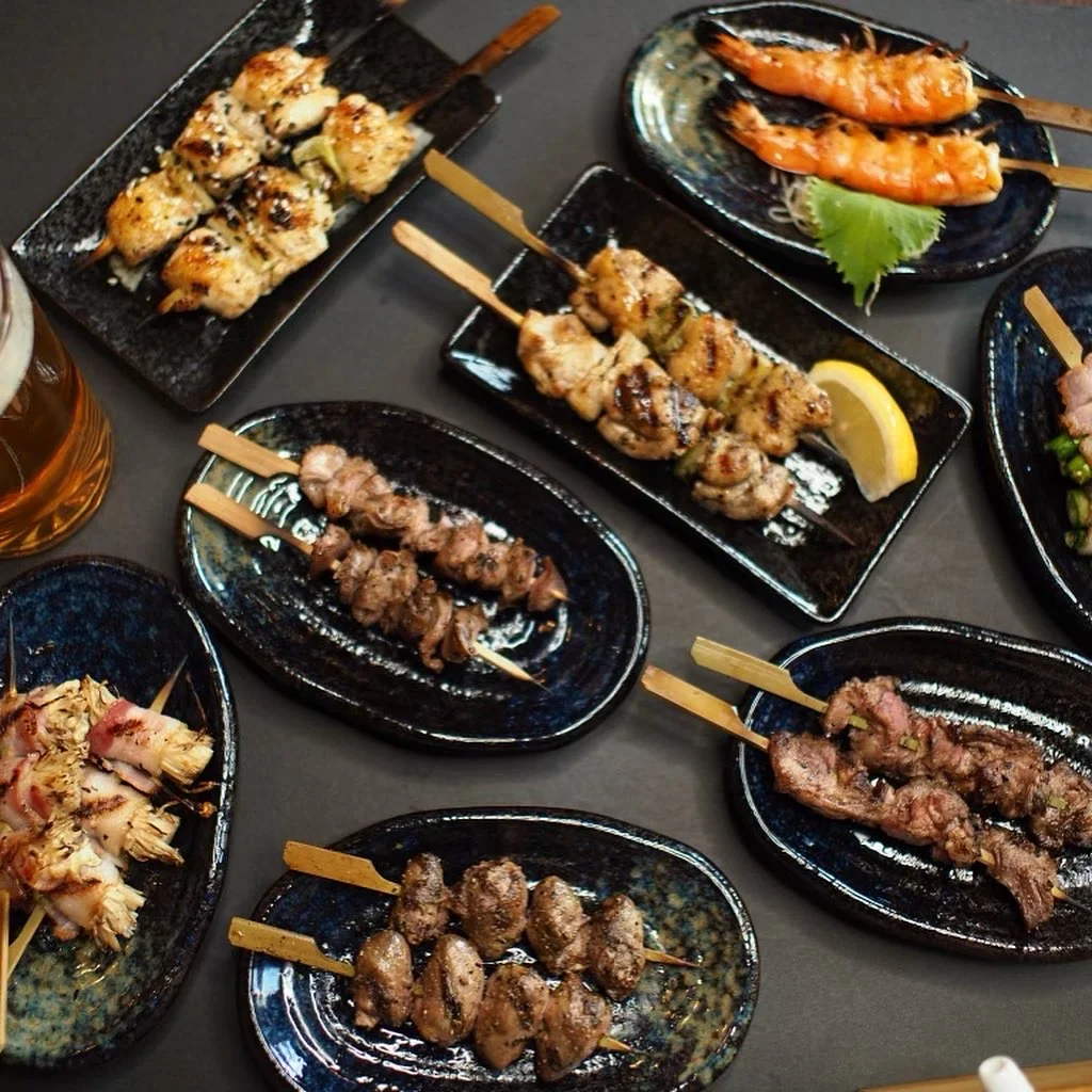An assortment of grilled skewers on dark ceramic plates with lemon wedges at Kinda Izakaya.